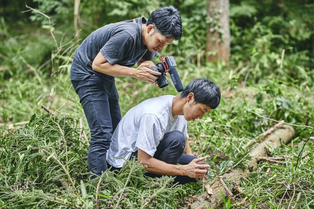 Tomomi wird von seinem Fotografen gefilmt, wie er mit einer Scheibenbremse einen Baumstamm schnitzt